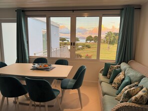 A room with a dining table and chairs, a sofa with cushions, and large windows offering a view of a building and a landscaped area.