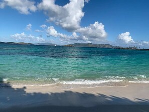 A beach with clear turquoise water, a sandy shore, and distant mountains under a partly cloudy sky.