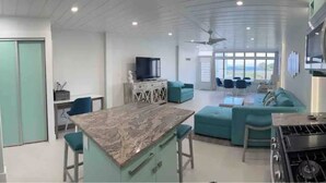 Et moderne køkken med en marmor bordplade, komfur og vask. Der er en teal farvet sofa og et fjernsyn i opholdsstuen.