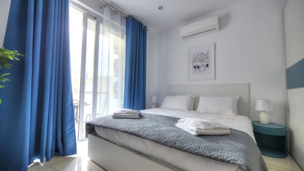 Moderni hotellihuone, jossa on suuri sänky, yöpöytä lampun kanssa ja ikkuna, jossa on siniset verhot.
