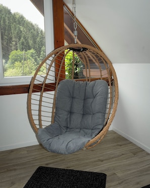 Een kamer met een hangstoel, een raam met uitzicht op groen en een houten vloer.