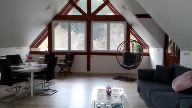 A spacious loft with a high ceiling, large windows, a dining area with a table and chairs, a hanging chair, and a sofa. - Vacation apartment Panoramablick (Oberharmersbach)