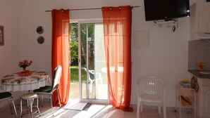A room with a dining table, chairs, a TV, and a sliding glass door with orange curtains. - Cozy studio in the garden with terrace (Lagoa / Porches)