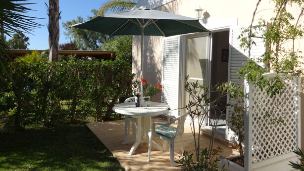 A patio with a table and chairs under a green umbrella, surrounded by greenery and a white fence. - Cozy studio in the garden with terrace (Lagoa / Porches)