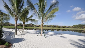 Una playa arenosa con palmeras y un lago tranquilo, rodeada de casas.