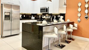 Une cuisine moderne avec des armoires blanches, un réfrigérateur en acier inoxydable, un four à micro-ondes et une machine à café. On y trouve des tabourets de bar et un grand îlot central doté d’un comptoir noir.