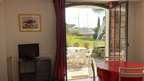A room with a television, a dining table with chairs, and a view of a garden through an open door. - Beautiful 3-person apartment facing the main beach (Saint-Georges-de-Didonne)