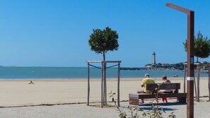 Une plage avec un banc en bois et un kiosque, un phare en arrière-plan et un ciel bleu dégagé.