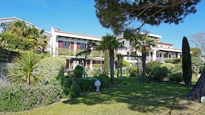 A building with multiple stories, a well-maintained garden with various plants and a lawn, and a clear blue sky. - Beautiful 3-person apartment facing the main beach (Saint-Georges-de-Didonne)