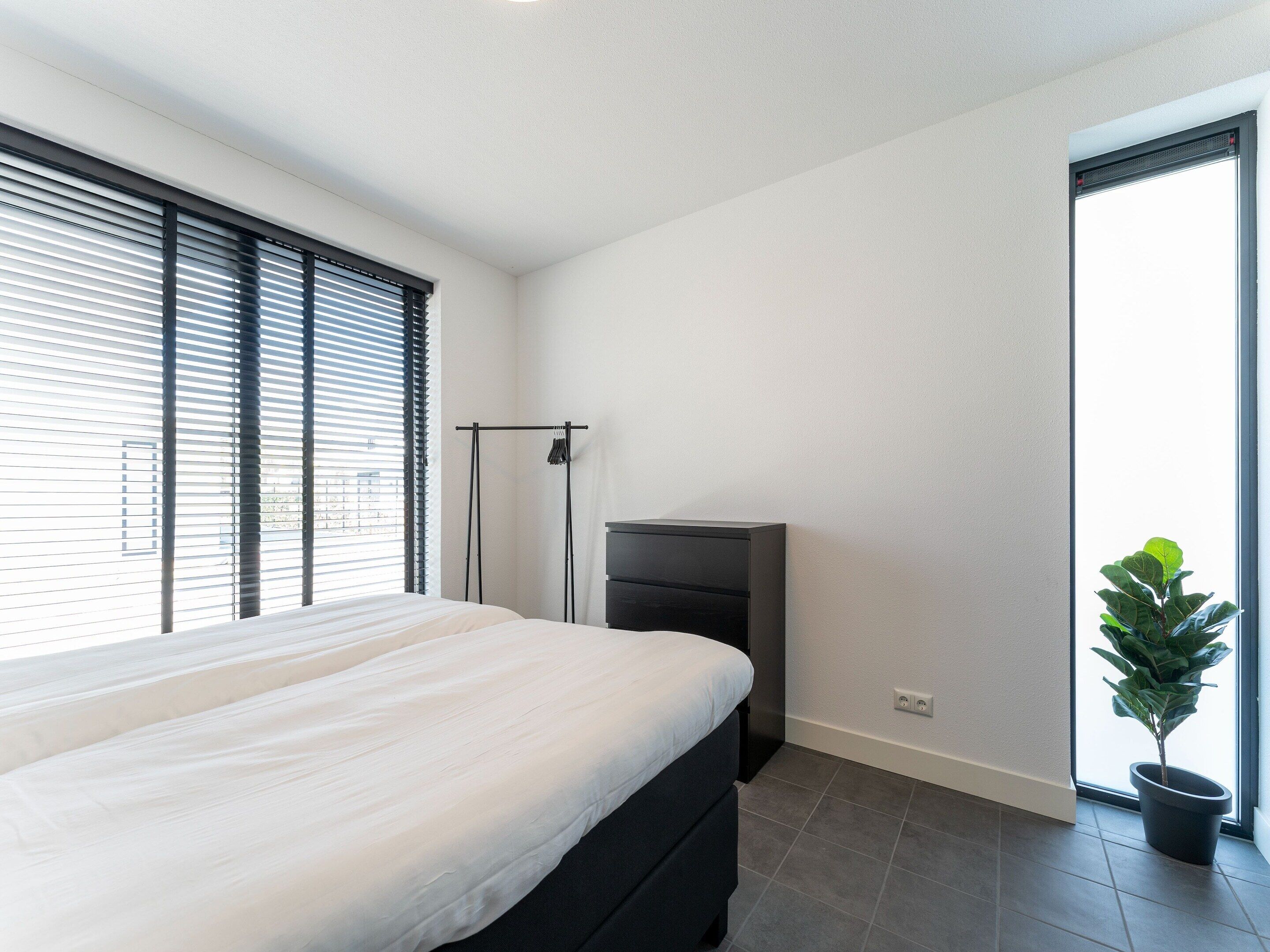 A modern bedroom with a bed, a black dresser, a floor lamp, and a potted plant by a large window.