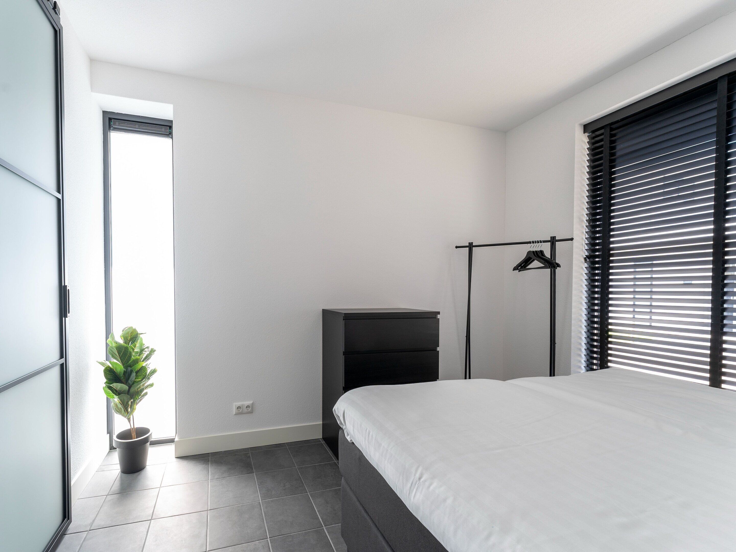 A modern bedroom with a bed, a small desk, a potted plant, and a window with blinds.