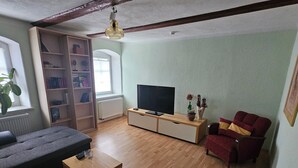 Un salon comprenant un canapé, une table basse, un téléviseur posé sur un meuble en bois, une bibliothèque et un fauteuil rouge.