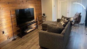 Een woonkamer met een flatscreen televisie op een standaard, een bank, een fauteuil en een lamp.