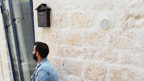 Un hombre sentado en un banco de hormigón frente a un edificio con pared de piedra, una puerta y una maceta con un cactus.