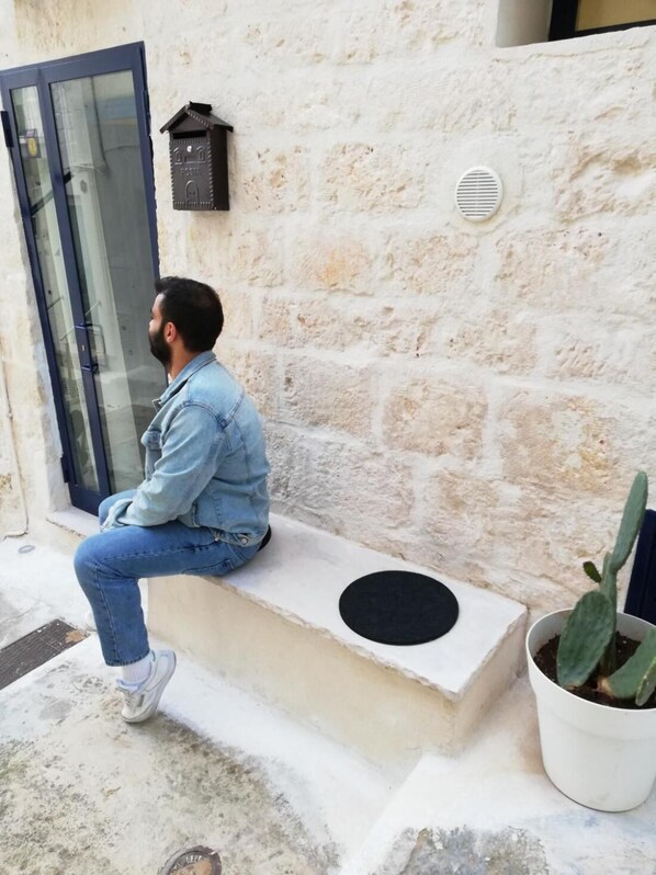 Un hombre sentado en un banco de hormigón frente a un edificio con pared de piedra, una puerta y una maceta con un cactus.