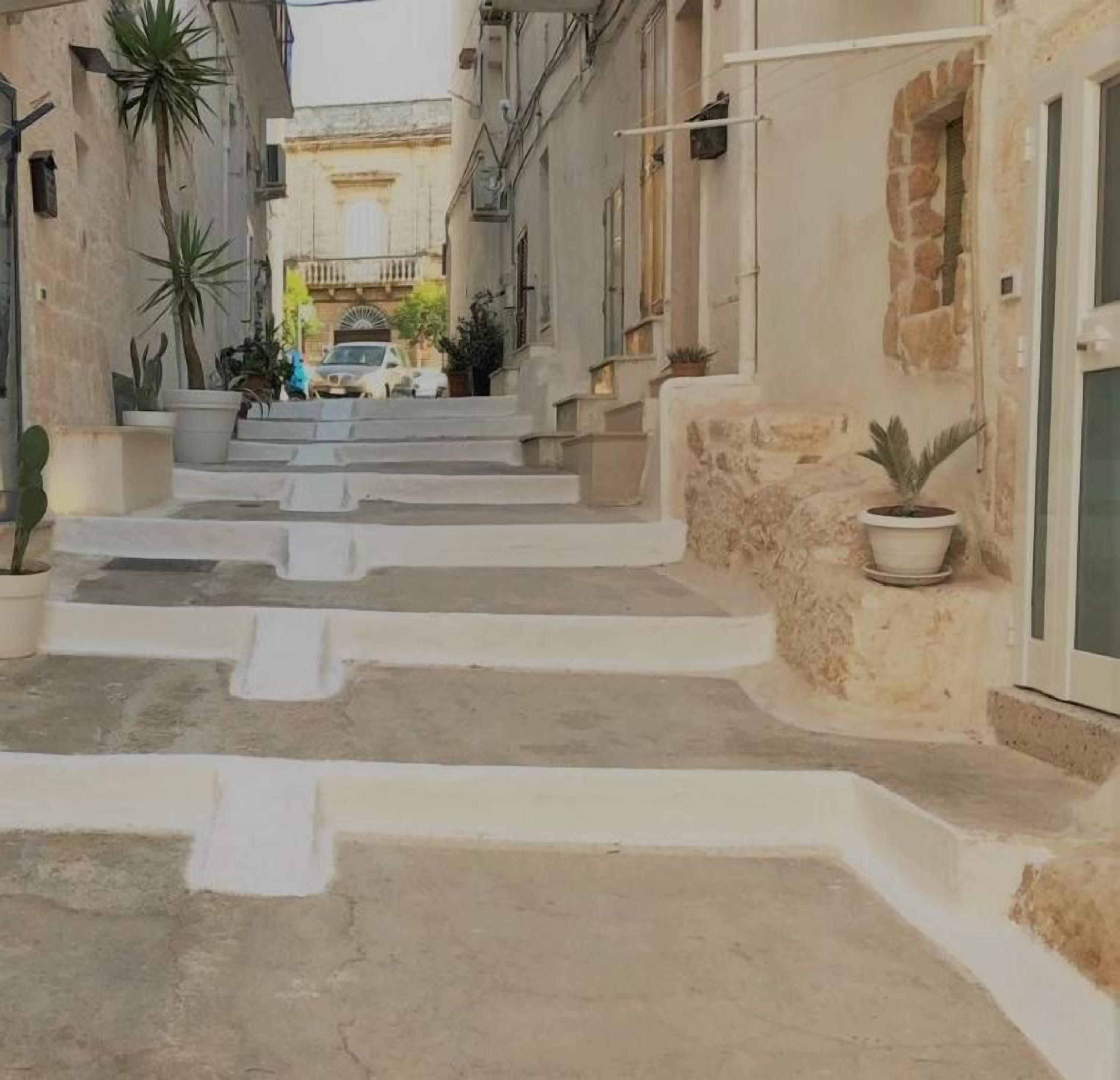 A narrow street with steps leading to a building. The building has a sign that reads "The Doors." There are potted plants on the steps and along the sides of the street.