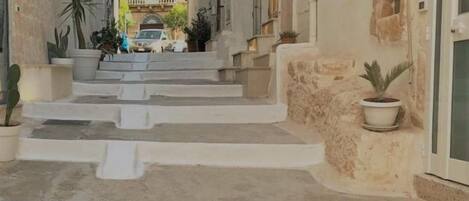 A narrow street with steps leading to a building. The building has a sign that reads "The Doors." There are potted plants on the steps and along the sides of the street.