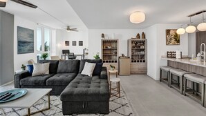 A modern living room with a grey sectional sofa, a dining area with a kitchen in the background, and a large window providing natural light.
