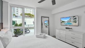 A modern hotel room with a large bed, a ceiling fan, a television, and a view of a swimming pool through a large window.