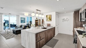 A modern kitchen with a marble island, wooden cabinets, and a built-in oven. There is a dining area with a large window overlooking the city.