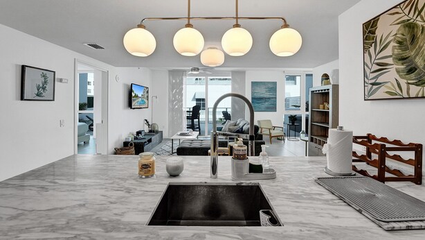 A modern kitchen with a marble countertop, a sink, and a built-in oven. There is a dining area with a table and chairs, and a large window providing a view of the outdoors.