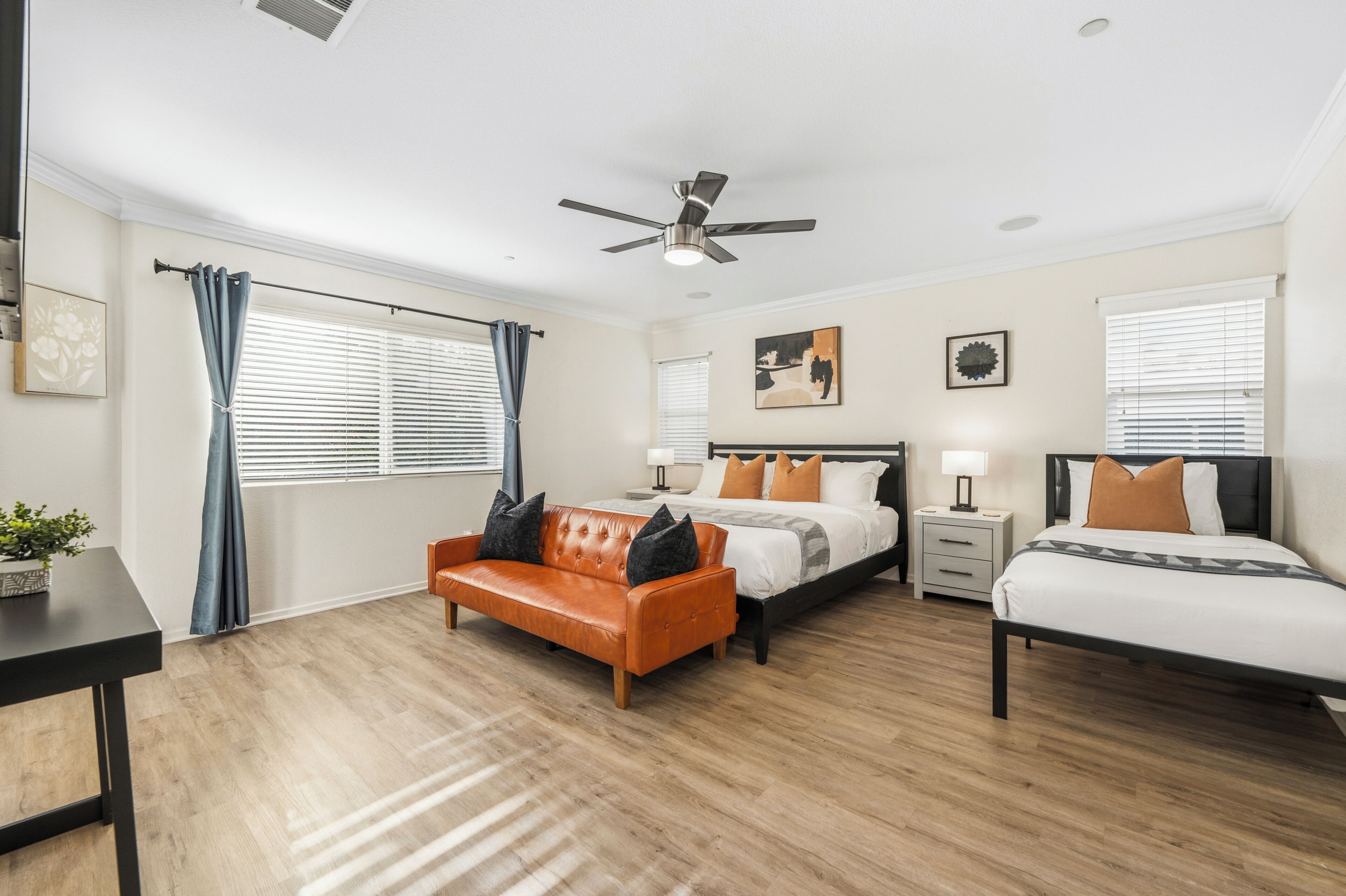 A bedroom with a bed, a sofa, a nightstand, a ceiling fan, and a window with blinds.