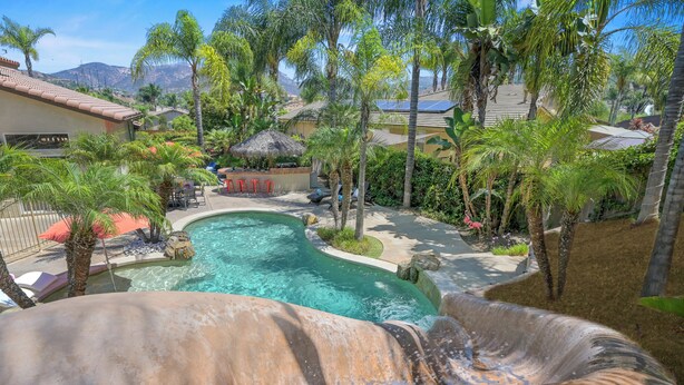 A pool with a waterfall feature, surrounded by palm trees and a thatched-roof structure. - Villa Paradiso (El Cajon)