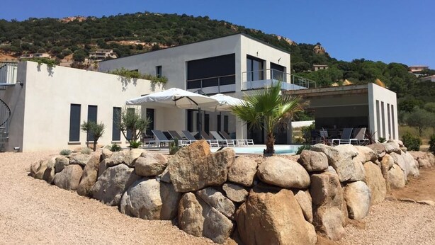Un edificio moderno con zona de piscina, grandes rocas al frente y una colina de fondo.