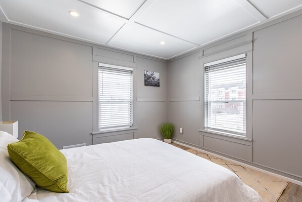 Une chambre à coucher avec un grand lit, deux fenêtres équipées de stores, une petite plante et un tableau accroché au mur.