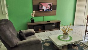 A living room with a grey sofa, a white coffee table with a flower arrangement, a TV on the wall, and a wooden cabinet. - POUSADA SERRANO (São Bento do Sapucaí)