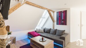 Une chambre à coucher de style loft moderne, avec un plafond en pente, un canapé gris, une table basse en bois et un tableau abstrait aux couleurs vives.