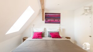 Une chambre avec un plafond en pente, un lit recouvert d’une couette grise, deux oreillers, un tableau au-dessus du lit et une tête de lit en bois.