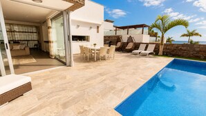 Un espace piscine extérieur moderne, doté d’un coin repas, de chaises longues, d’une terrasse en pierre et offrant une vue sur l’océan.