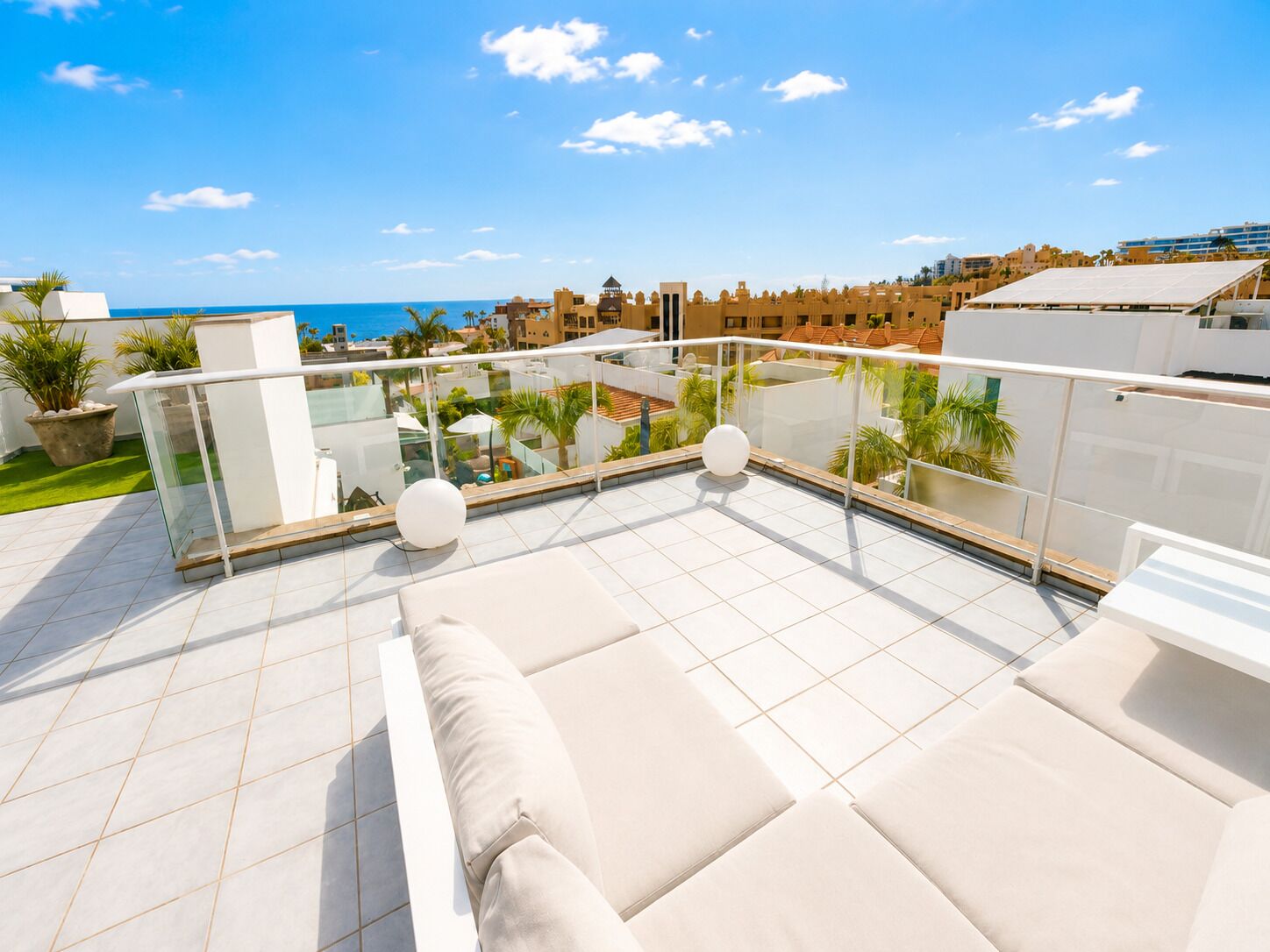 A rooftop terrace with a glass balustrade, outdoor seating, and a view of the sea and buildings.