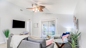 Ein modernes Wohnzimmer mit einem grauen Sofa, einem Flachbildfernseher, einem Deckenventilator und einer Glastür, die auf einen Balkon führt.