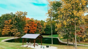 A gazebo with picnic tables and a bench, surrounded by a pond and trees with autumn foliage.