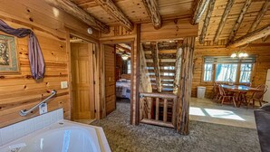 A log cabin interior with a bathroom featuring a bathtub, a wooden staircase, and a dining area with a table and chairs. - Cozy Pine at Tranquil Acres (Millersburg)