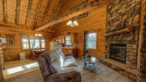 A log cabin interior with a stone fireplace, a sofa, a dining area, and a kitchen. - Cozy Pine at Tranquil Acres (Millersburg)