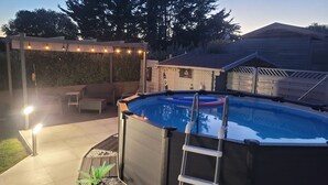 An outdoor swimming pool with a ladder, surrounded by a patio area with a pergola and string lights. - Family house, walking distance to the beach, near St Malo (Saint-Coulomb)
