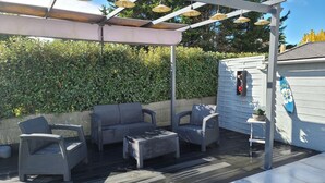 An outdoor patio with wicker furniture, a pergola with hanging lights, a hedge, and a small house with a blue sign. - Family house, walking distance to the beach, near St Malo (Saint-Coulomb)