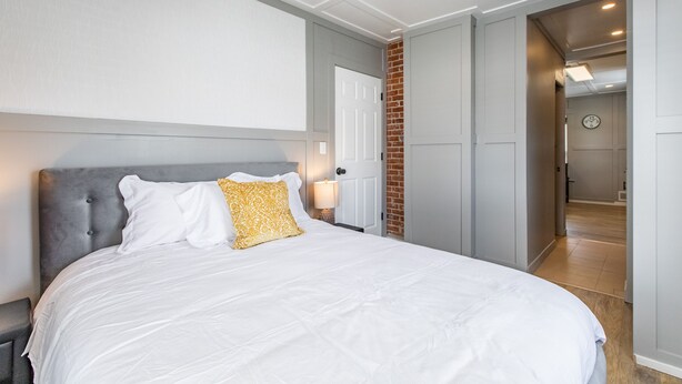 A bedroom with a large bed, a grey headboard, a white wall, and a door leading to another room.