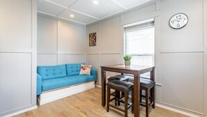 A room with a blue sofa, a wooden table with two stools, a clock on the wall, and a window with blinds.