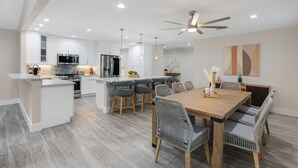 Une cuisine moderne avec des meubles blancs, un îlot central et un coin repas comprenant une table en bois et des chaises en osier.
