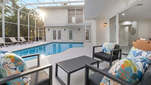 Un espace piscine intérieur moderne, doté de chaises longues et d’une table, de grandes fenêtres et d’un escalier menant à un niveau supérieur.