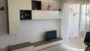 A modern living room with a flat-screen TV on a wooden console, a white shelving unit with decorative items, and a large window with sheer curtains.