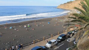 A beach with people sunbathing and swimming, a parking area with cars, and a cliff overlooking the ocean.