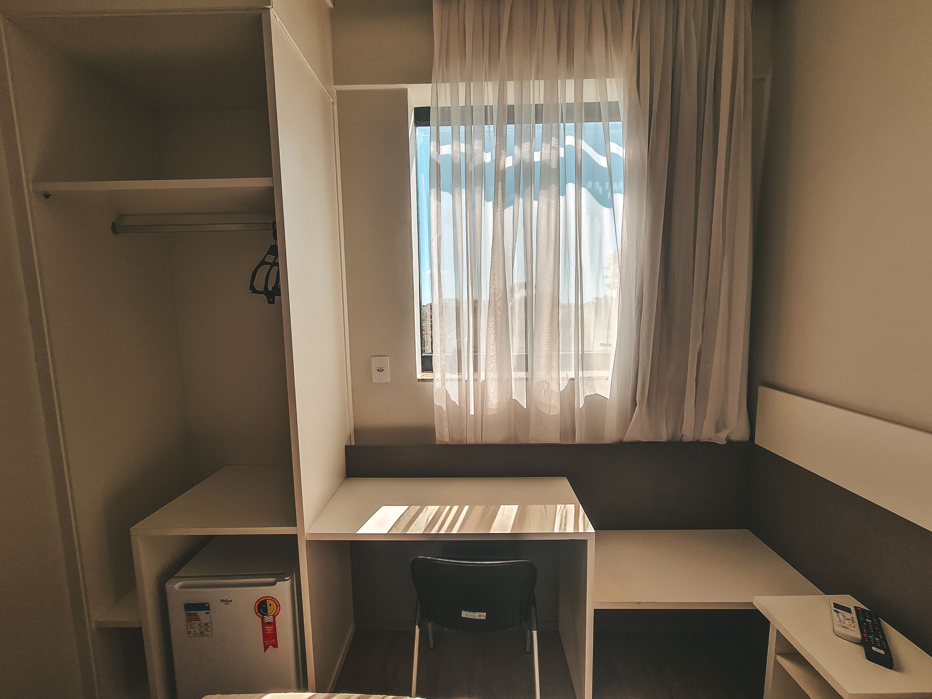 A compact room with a bed, a small desk, a chair, a mini-fridge, and a window with sheer curtains.