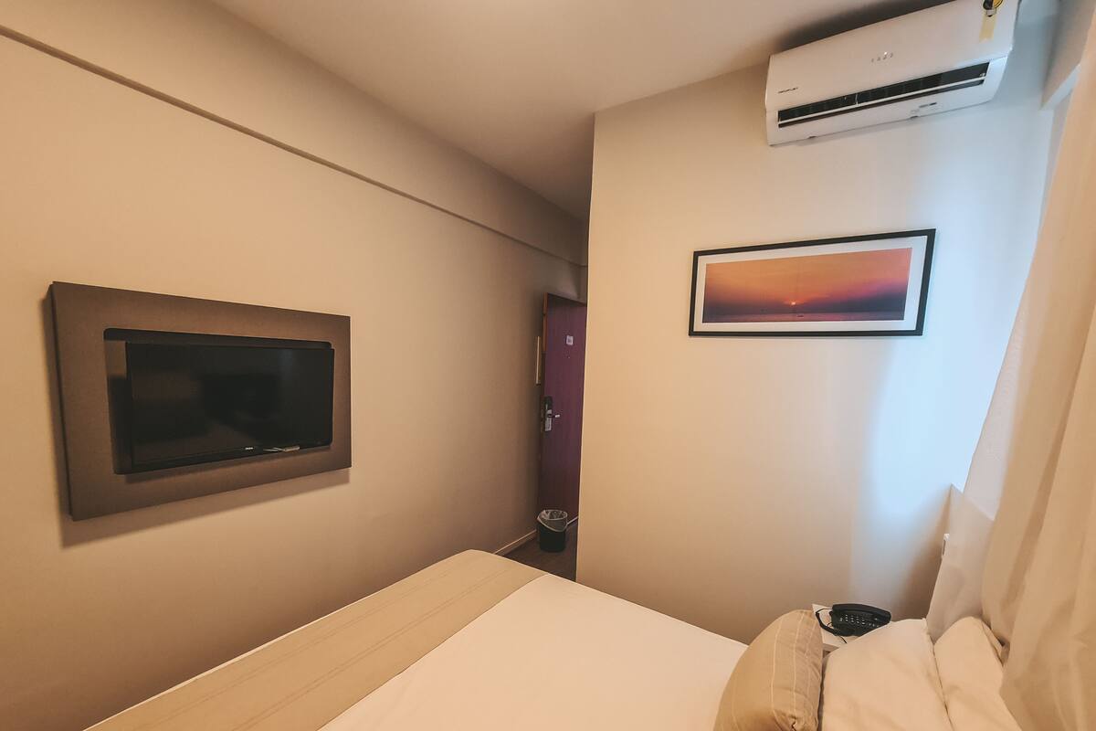 A hotel room with a bed, a mounted television, an air conditioning unit, and a framed picture on the wall.