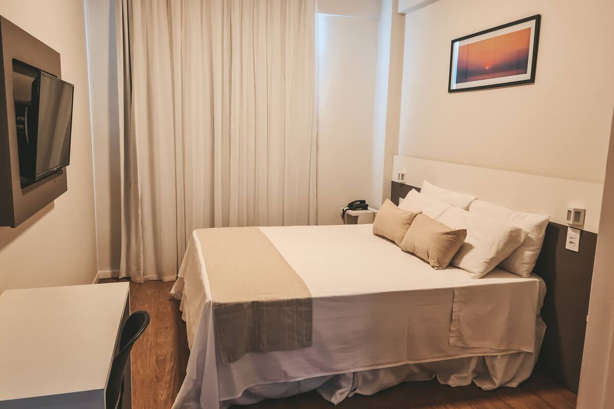 A hotel room with a bed, a desk, a chair, a TV, a nightstand, and a picture on the wall.