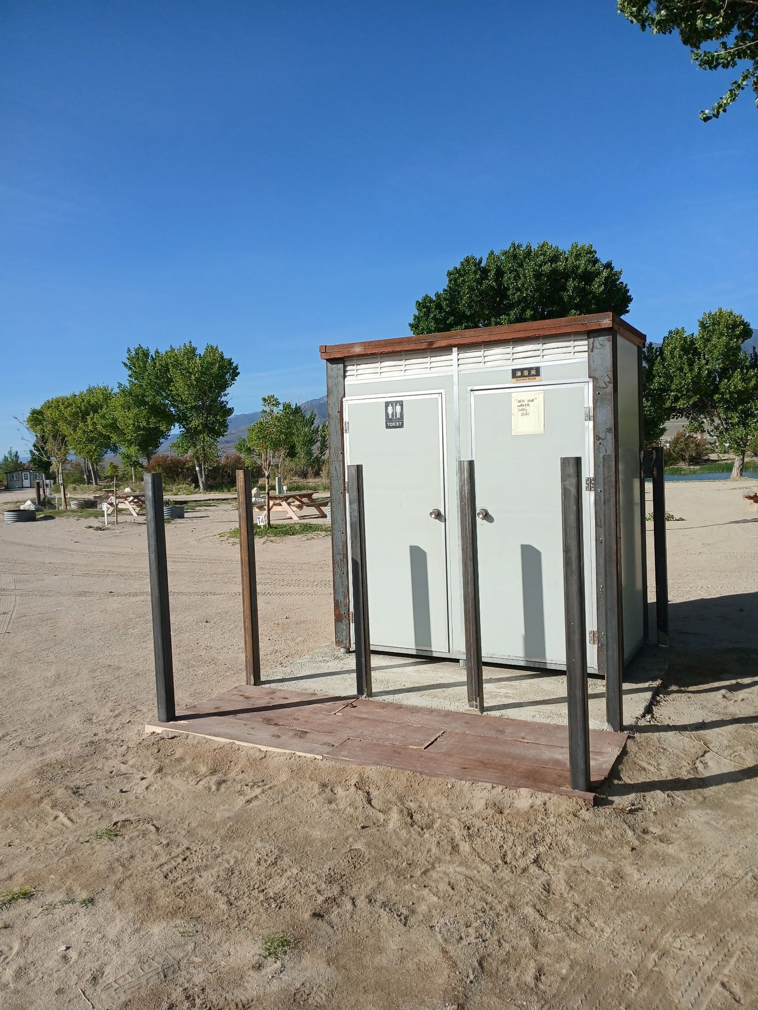 Unit portàtil de lavabo amb indicació que és per homes i dones, situada en una zona sorrenca amb arbres i un cel clar al fons.
